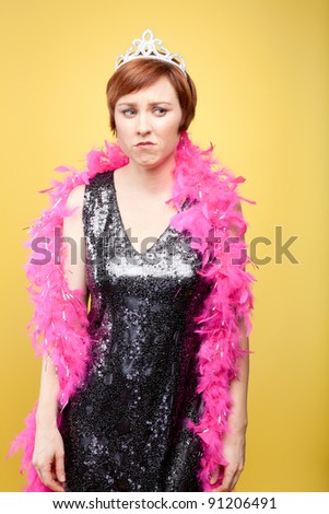 Unhappy  woman wearing an evening gown, plastic tiara and feather boa. Photographed with studio lighting in front of a yellow backdrop.
