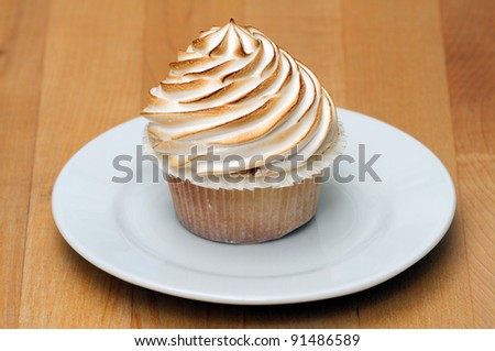 A cupcake with lots of meringue on top, browned in an oven, plated and placed on a maple tabletop.