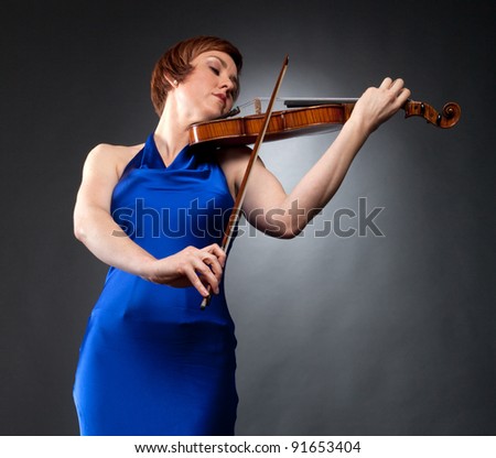 A woman in a blue evening gown plays the violin. Photographed with studio lighting in front of a gray backdrop.