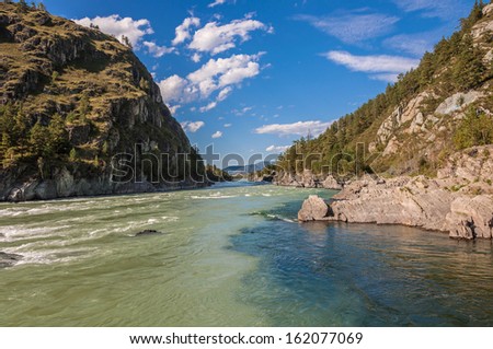 Confluence of two rivers in the mountains. In one of the rivers dark turquoise water. The rivers are flowing in the mountains, on the banks of the spruce. Summer sunny day with clouds.
