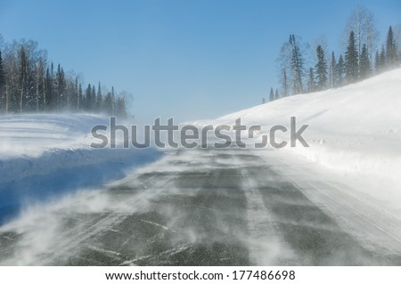 Drifting snow on winter road with snowdrifts on the roadside in the snowstorm on a sunny day.