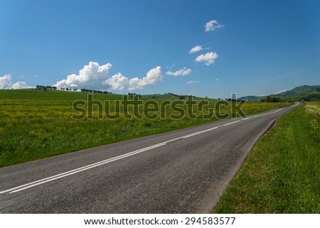 Scenic view from the asphalt road among meadows with yellow flowers on a background of mountains and blue sky with clouds