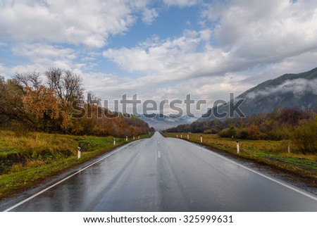 Bright picturesque view from the asphalt road, wet after the rain, mountains, fog, golden trees and grass against a blue sky with clouds