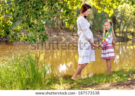 portrait of pregnant woman, ukrainian style, white dress, with daughterin park, near water, river