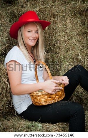 Country girl with red hat and basket to collect eggs sitting on a hay bale