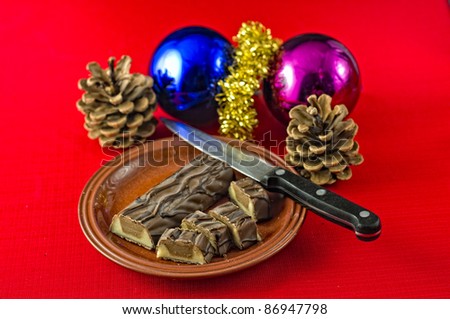 Christmas treat, chocolate covered marzipan and nougat, bar, partly cut on plate on red tablecloth. Christmas decorations on table. Selective focus on chocolate bar.