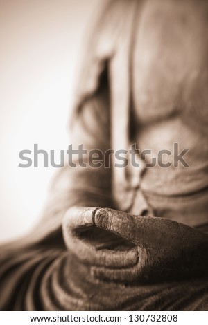Sepia photograph of antique carving of Buddha with hands folded in prayer, very shallow depth of field, Lensbaby image; focus on folded hand