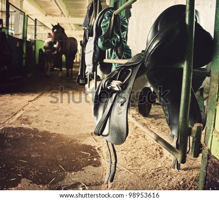 forefront of the saddle, harness, in the background a woman cleans horse