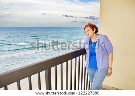 A pleasant looking, red haired, senior woman gazes dreamily off her balcony at the ocean waves feeling the cool ocean breeze on her face.