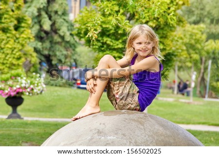 A young girl sits on a man made rock to take a break at an outdoor park just watching the world pass by while a light summer breeze rustles her hair.