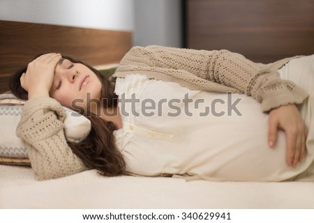 Beautiful young pregnant woman feeling head ache and laying on the bad in the room with modern furniture