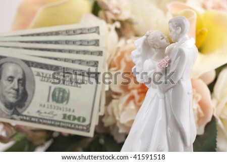 A wedding cake topper showing a groom and bride with one hundred dollars fanned out in the background with flowers