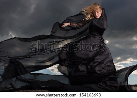 Wind blowing blond hair and long black dress of the young girl standing on the mount