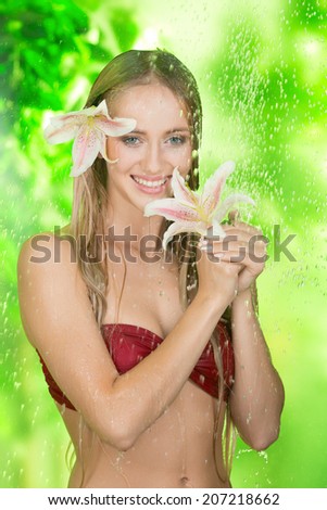 Cute smiling blonde girl with a flower in her hair and hands enjoys fresh shower outdoors
