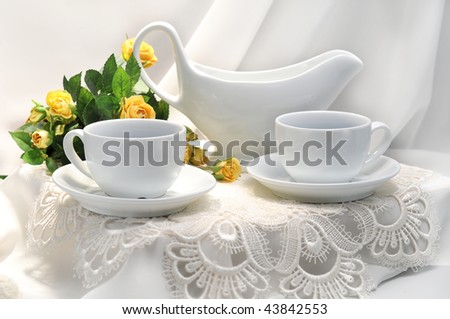 The wall in the background is a light off-white. Bowl on table is empty, suggesting the food has yet to arrive.White cups on a white table, a white lacy napkin, a green branch with yellow small roses.