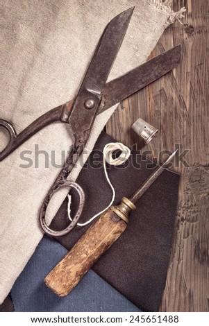 Vintage scissors, awl and thimble with leather and cloth on old wooden background