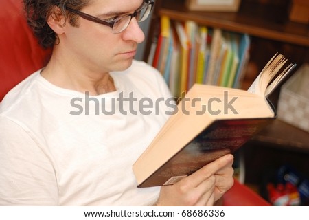 A Close Up of a Man Reading a Book, While Sitting on a Red Chair with a Bookcase in the Background