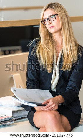 Portrait of an attractive businesswoman sitting on an office table working on a document or contract, thoughtful look into nowhere with her legs visible under a short skirt.