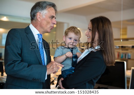 Reconciliation of family and work life: Attractive brunette woman in business attire proudly carrying a small ,boy in her arm in office environment discussing with her boss who seems not amused