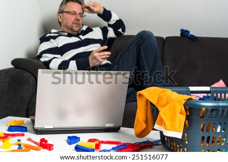 reconciliation of work and family life: Portrait of a middle aged man sitting in front of computer with toys and baby clothes on sofa, doing a phonecall