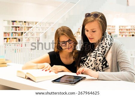 Two attractive female students are stuyding at the public library using a digital tablet and books