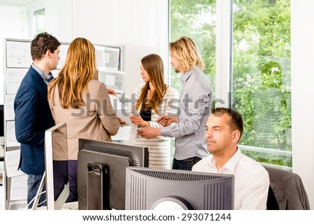 Businesspeople working in the office - some of them looking at bulletin board and discussing designs pinned at it, another guy in the front is working with several computer screens. Mixed caucasian