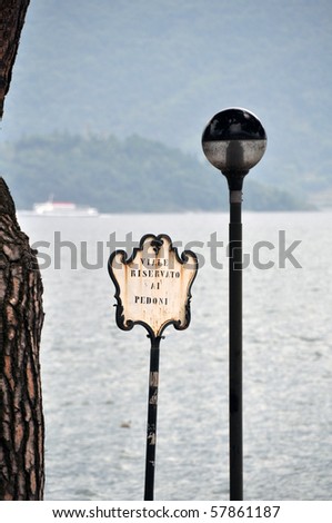 Idyllic Italian waterfront in Domaso at Lake Como with old streetlight, pedestrian area sign and pine tree, passenger boat in the background