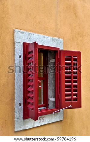 Red Window Shutters opened on a bright frame above a terracotta colored wall. The shutters unveil an also red lattice window. Underneath the window the house number 55 is painted.