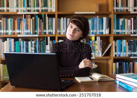 Student girl sitting with books and laptop computer in college library. Looking to camera. Selective focus on face.