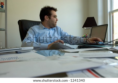A businessman in a blue shirt working on a laptop at his desk with cluttered papers.