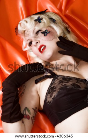 A blond woman with a black vintage bra, black gloves, and black hair net laying on a red silk cloth.