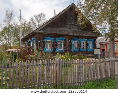 Brown wooden house with blue fretted window cases and palisade before against cloudy sky background. Nizhegorodsky region, Russia