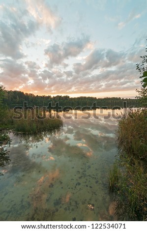 Sunset over Svetloyar Lake with bush in foreground and forest along bank reflecting in calm water