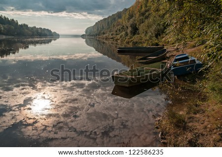 Several green wooden boats sunken and laid up on riverbank against wide panorama of calm Vetluga river with sun reflection. Nizhegorodsky region, Russia.