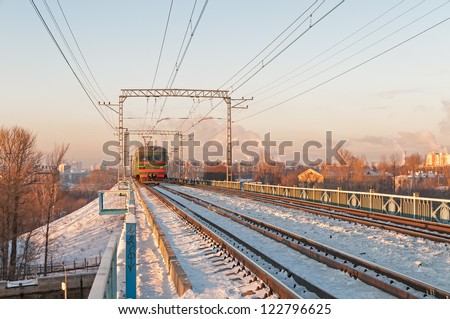 Green suburban electric train moves towards on snow-bound railroad vanishing in horizon before sunset light against skyline background. Moscow, Russia.