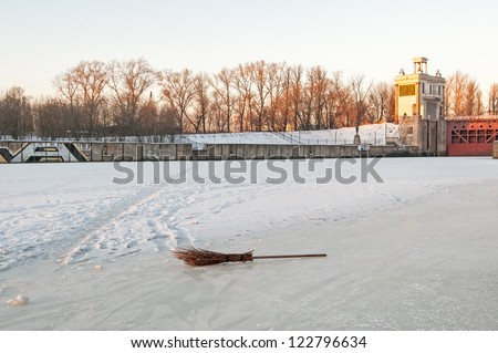 Broom on ice field and numerous footprints and beat path in snow against concrete wall and closed shipping lock of frozen canal background. Moscow, Russia.