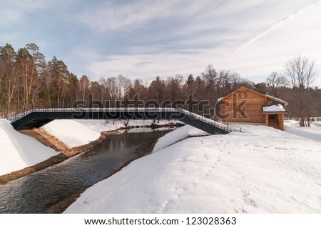 Black metal bridge over river with snow-covered banks against brown log cabin and forest background. Nizhegorodsky region, Russia.