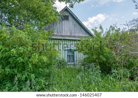 Old abandoned green wooden house with plantband on window among wild tall weeds and trees. Bolshaya Doroga village, Tambovsky region, Russia.