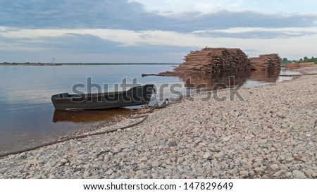 Wooden boat and timber store sinks in water along riverside against cloudy sky background. Severnaya Dvina River