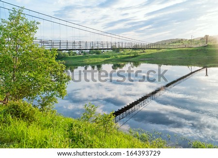 Old lengthy hanging wooden footbridge with rails over river. Arkhangelsky region, Russia.