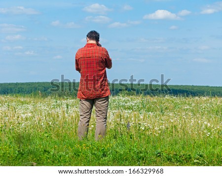 Tall man in bright check shirt stands back before beautiful sunny blossoming meadow with forest in background. Kaluzhsky region, Russia.