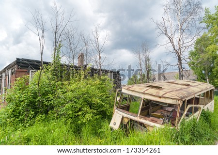 Crashed rusty bus in grass on damaged wooden house background.