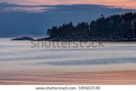 View on Onega Lake and cape with lighthouse among trees against evening glow background at midnight sun. Besov Nos cape, Karelia Republic, Russia