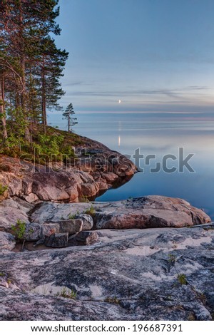 View on Onega Lake granite shore and evening glowing with crescent reflecting in calm water at midnight sun. Besov Nos cape, Karelia Republic, Russia.