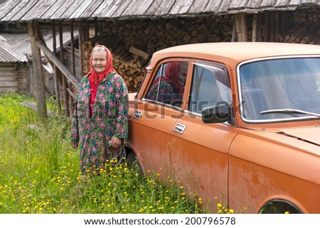 KVAZENGA, ARKHANGELSKY REGION/RUSSIA - JUNE 23: Old woman in red headscarf stands near orange car before blurred stack of firewood background on June 23, 2010 in Kvazenga.
