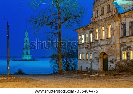 Winter view on old mansion on riverbank with famous drowned belfry of St. Nicholas cathedral (1800) standing in middle of frozen reservoir in background at night. Kalyazin, Tverskaya region, Russia.