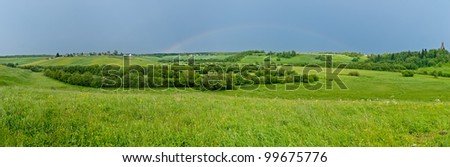 Wide panorama of hilly landscape with full rainbow in the sky, villages and church in background.