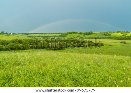 Panorama of hilly landscape with full rainbow in the sky against village in background.