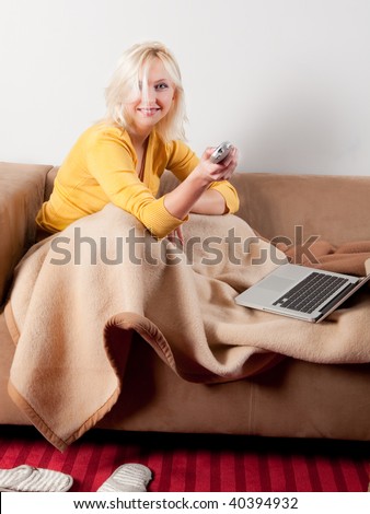 young woman wrapped in blanket relaxing on sofa, whatching TV and having laptop