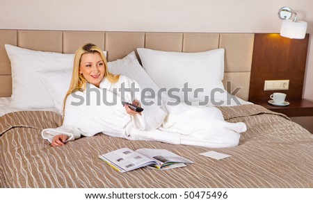 An attractive woman in a bathrobe lies on a bed. Watching TV. Horizontal shot.